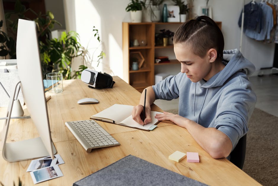 A teenage boy focuses on his studies at home, writing in a notebook during an online class.