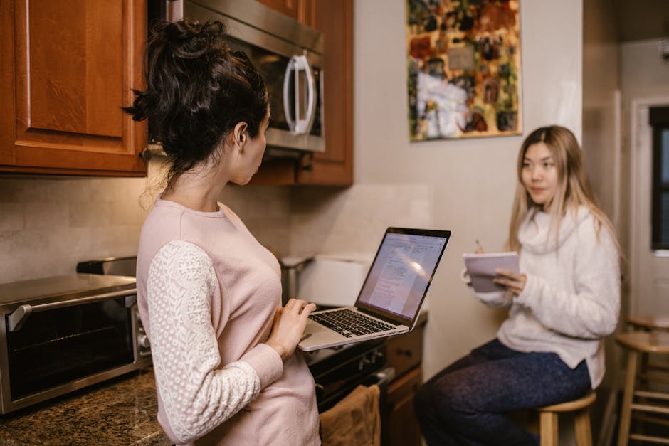 Two women collaborate on laptops and notes in a cozy home kitchen setting.